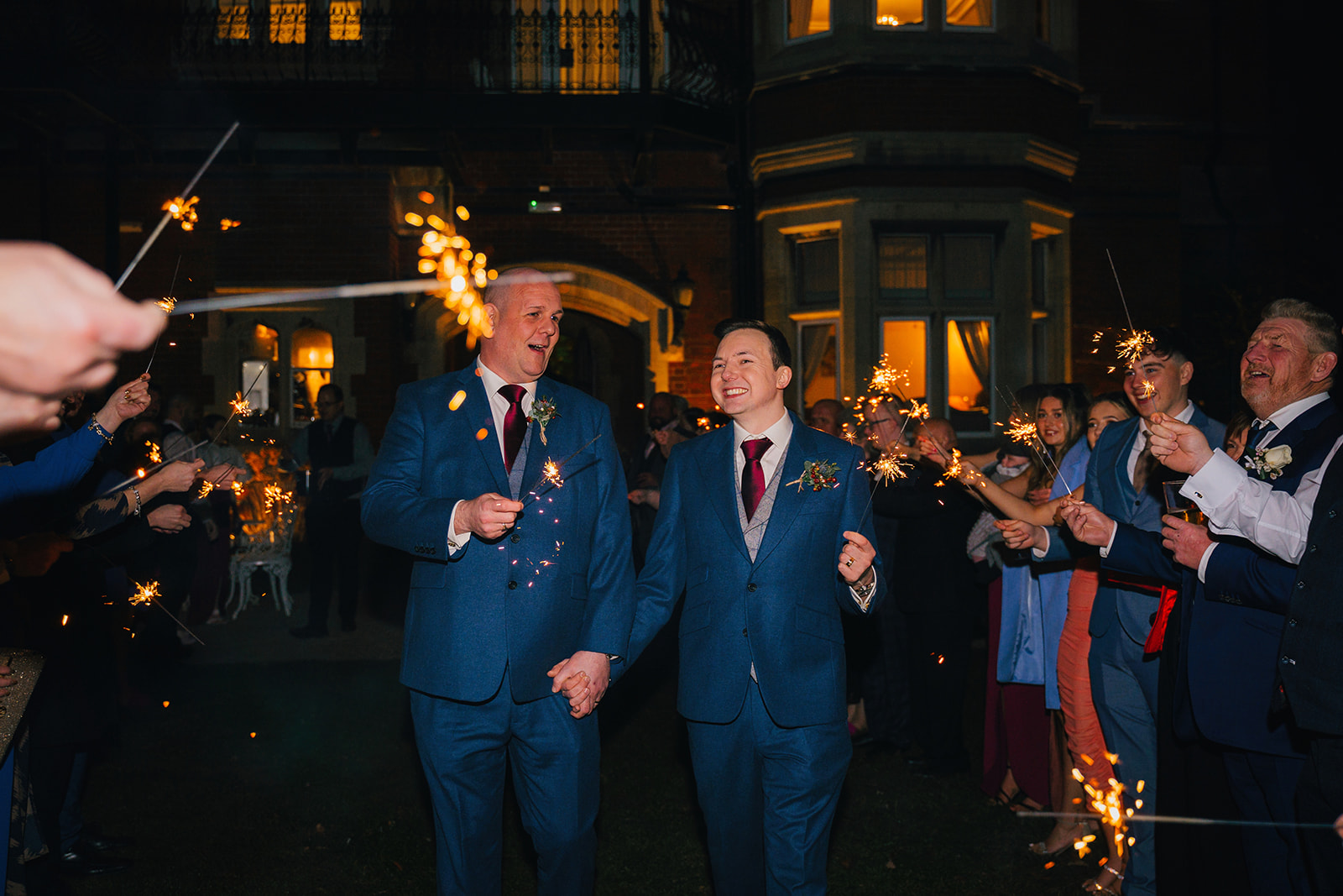 guests line up either side as the two grooms walk hand in hand down an aisle, waving sparklers
