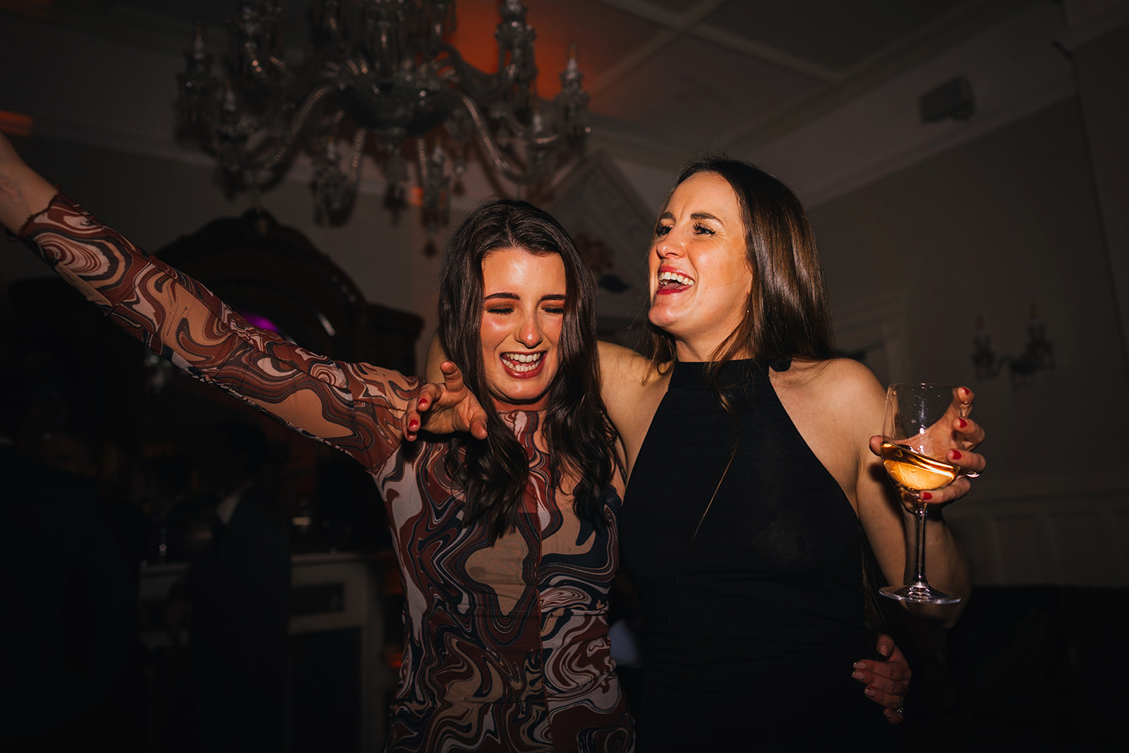 two women throw their arms in the air as they hold their wine glasses and dance