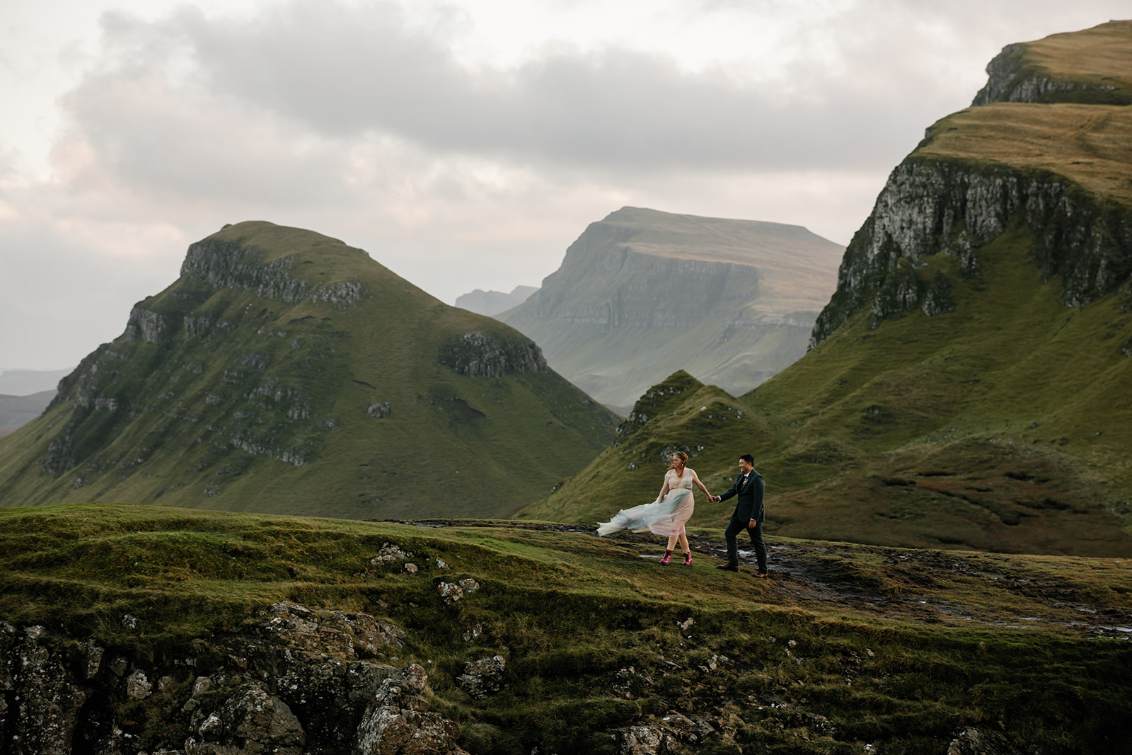 Mellie and Andrew stand at the edge of a rocky cliff overlooking the breathtaking Quiraing landscape on the Isle of Skye
