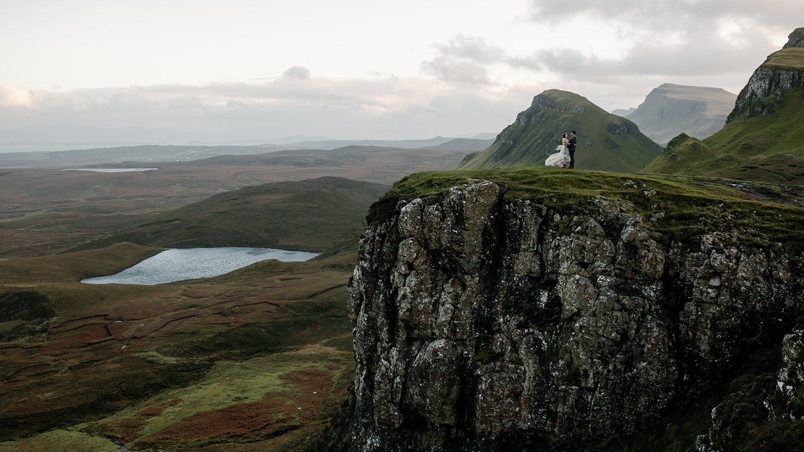 Mellie and Andrew stand at the edge of a rocky cliff overlooking the breathtaking Quiraing landscape on the Isle of Skye
