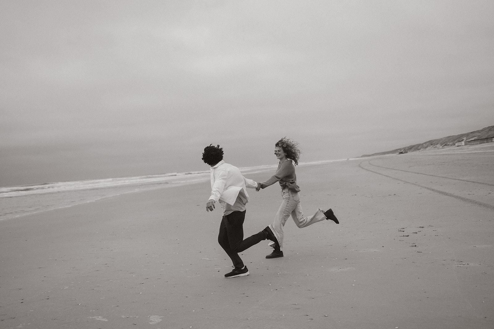 Couple runs direction ocean while holding hands. It's windy.