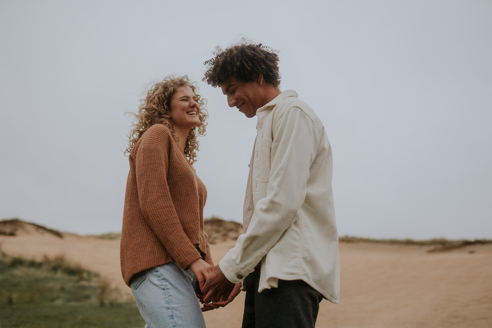Couple standing in front of each other holding hands softly. In the background there is a sand dune. 