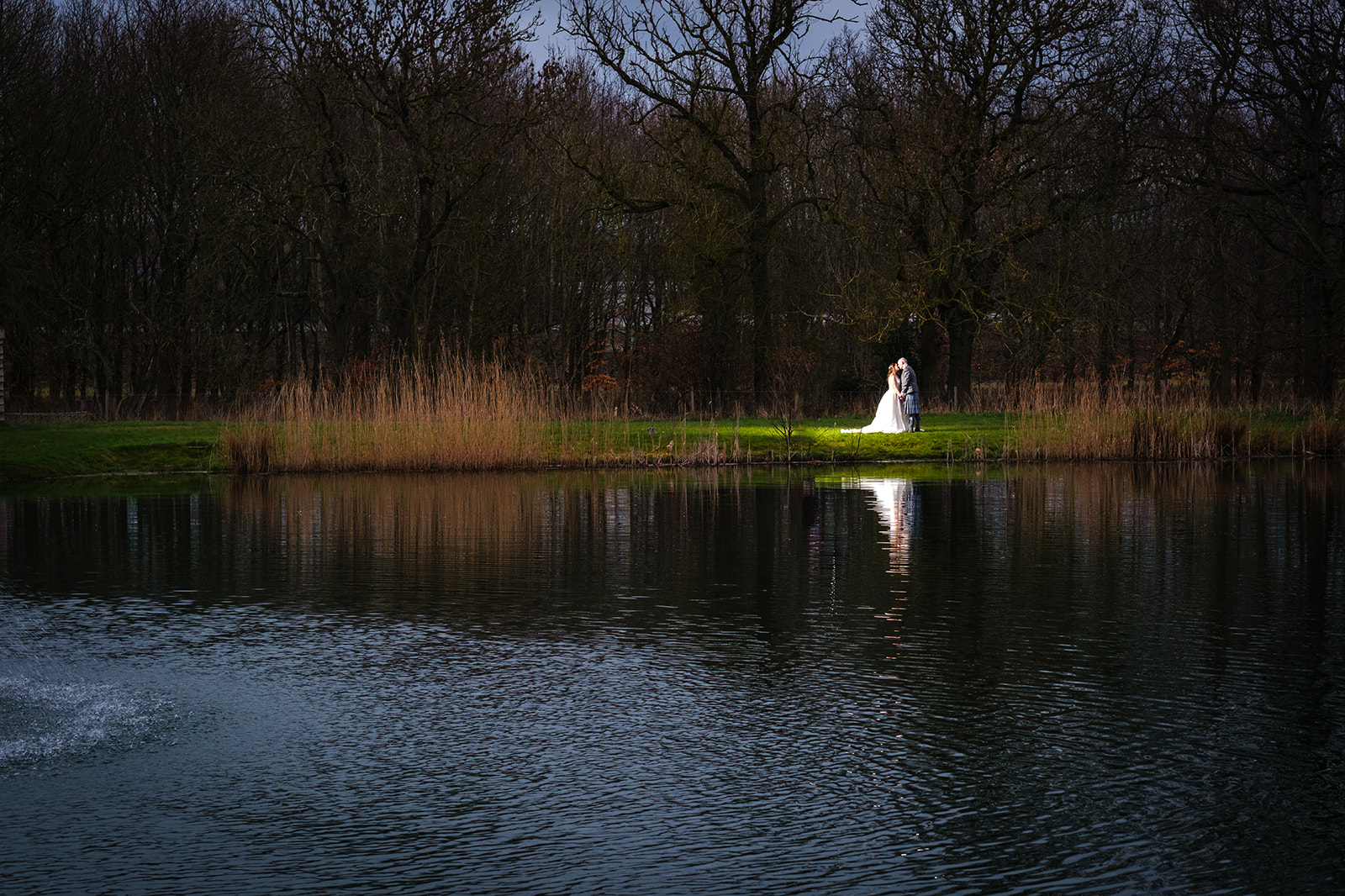 Married couple standing on one side of the lake being illuminated by flash on the brides wedding dress.