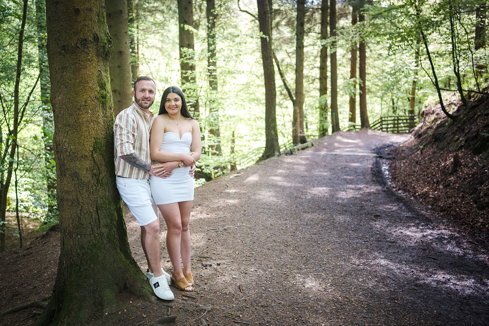 Engaged couple leaning against a tree along a path through a section of woodland