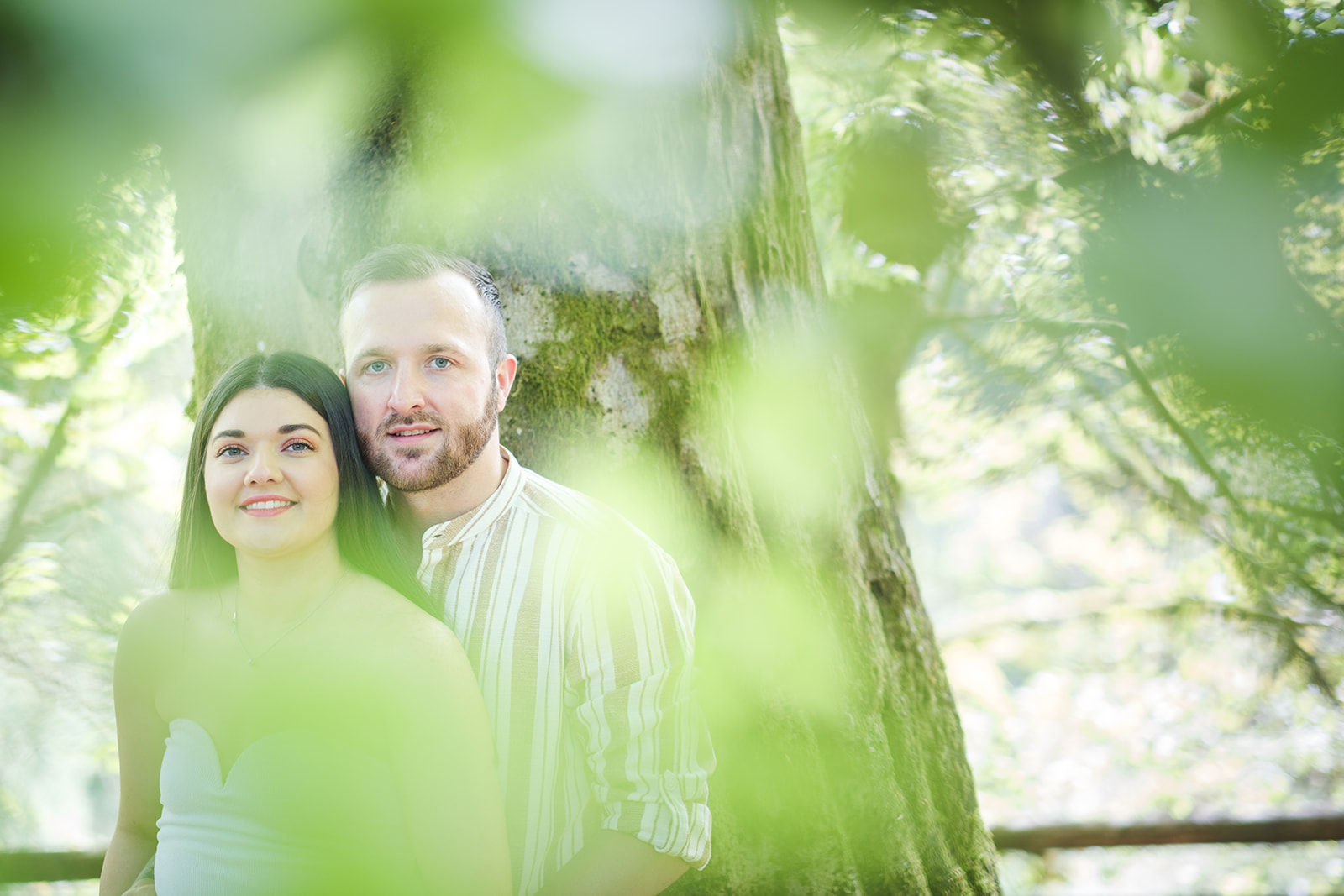 Engaged couple leaning on a tree whilst photographer take a photo through the leaves