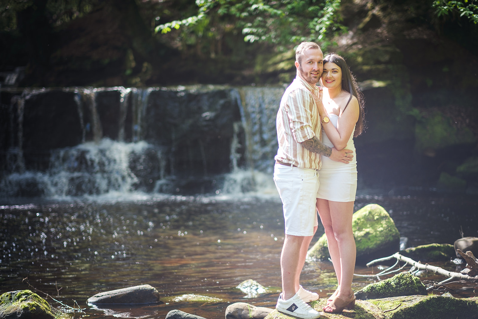 Engaged couple standing in front of a small waterfall at Calderglen Park in East Kilbride