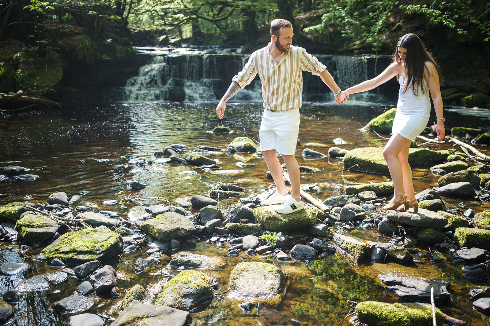 Engaged couple walking across make shift stepping stones across the river whilst holding hands