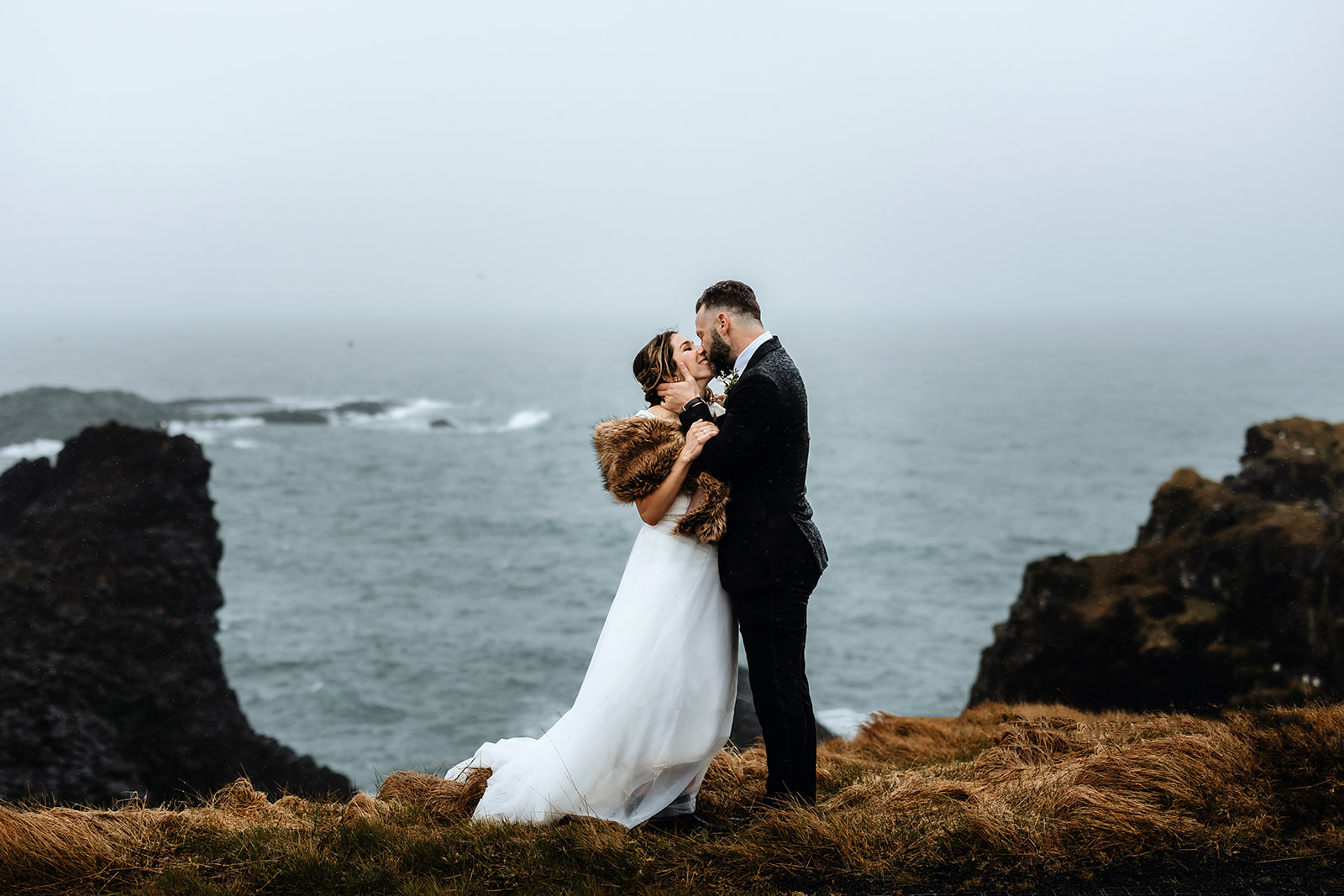 Married couple is hugging in Arnarstapi in front of the ocean and cliffs in Iceland