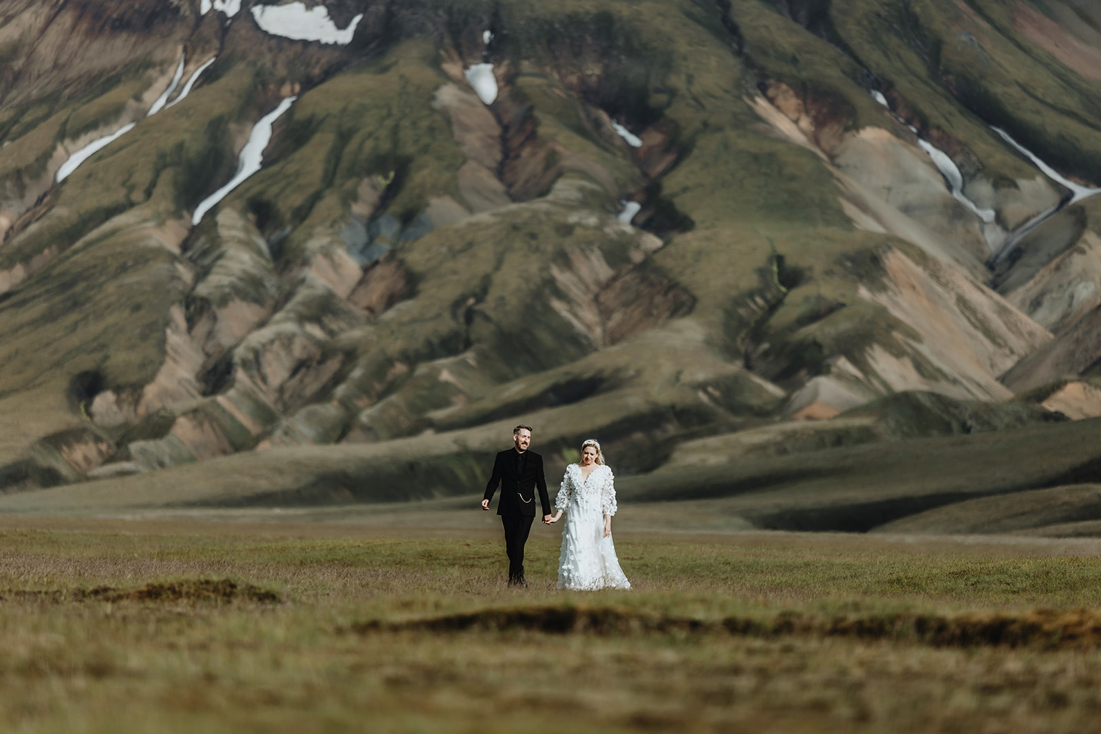 Newlywed couple holding hands and walking through the Icelandic highlands, with rolling hills and a serene landscape. 
