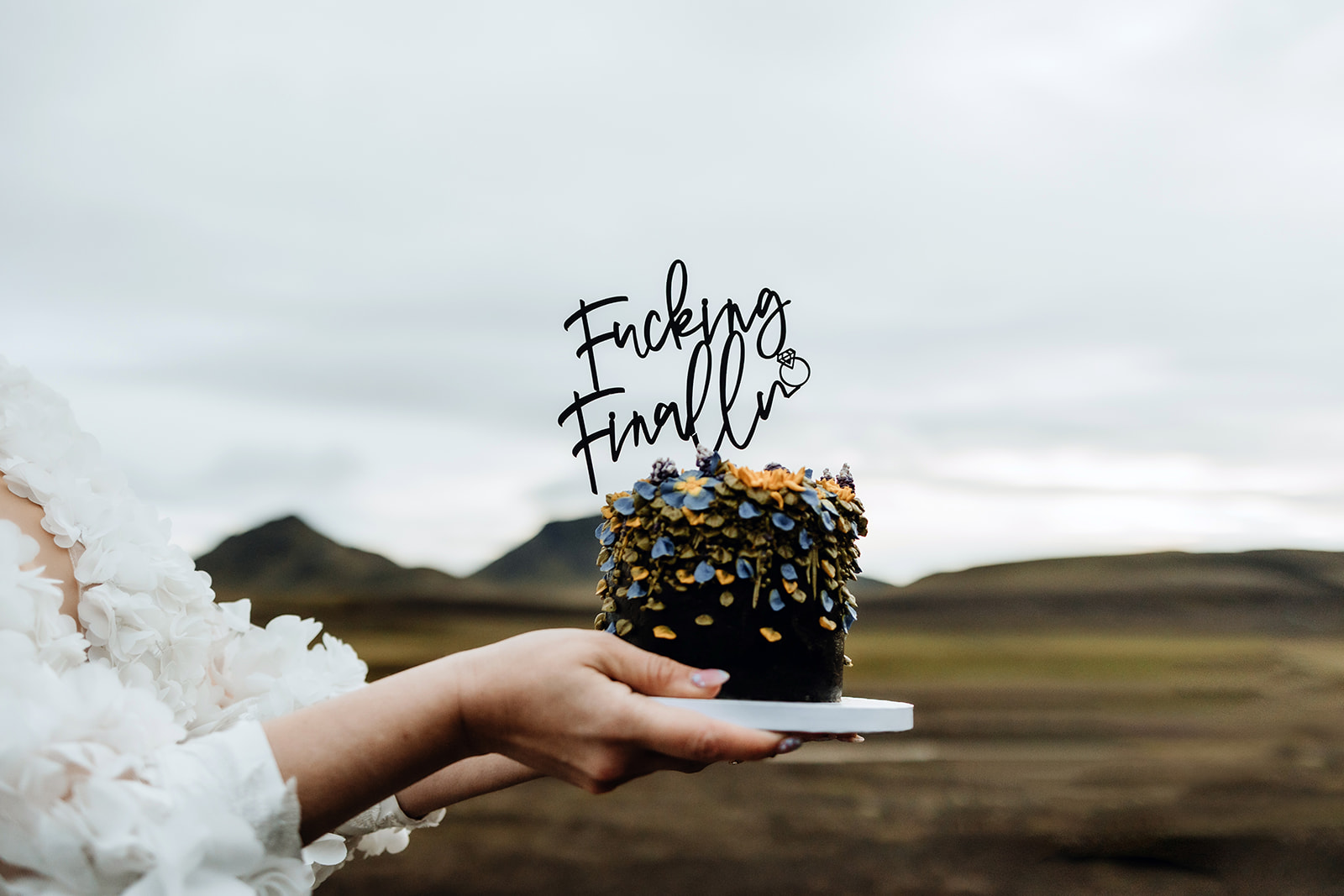 A bride holds a wedding cake that says 'fucking finally'. 