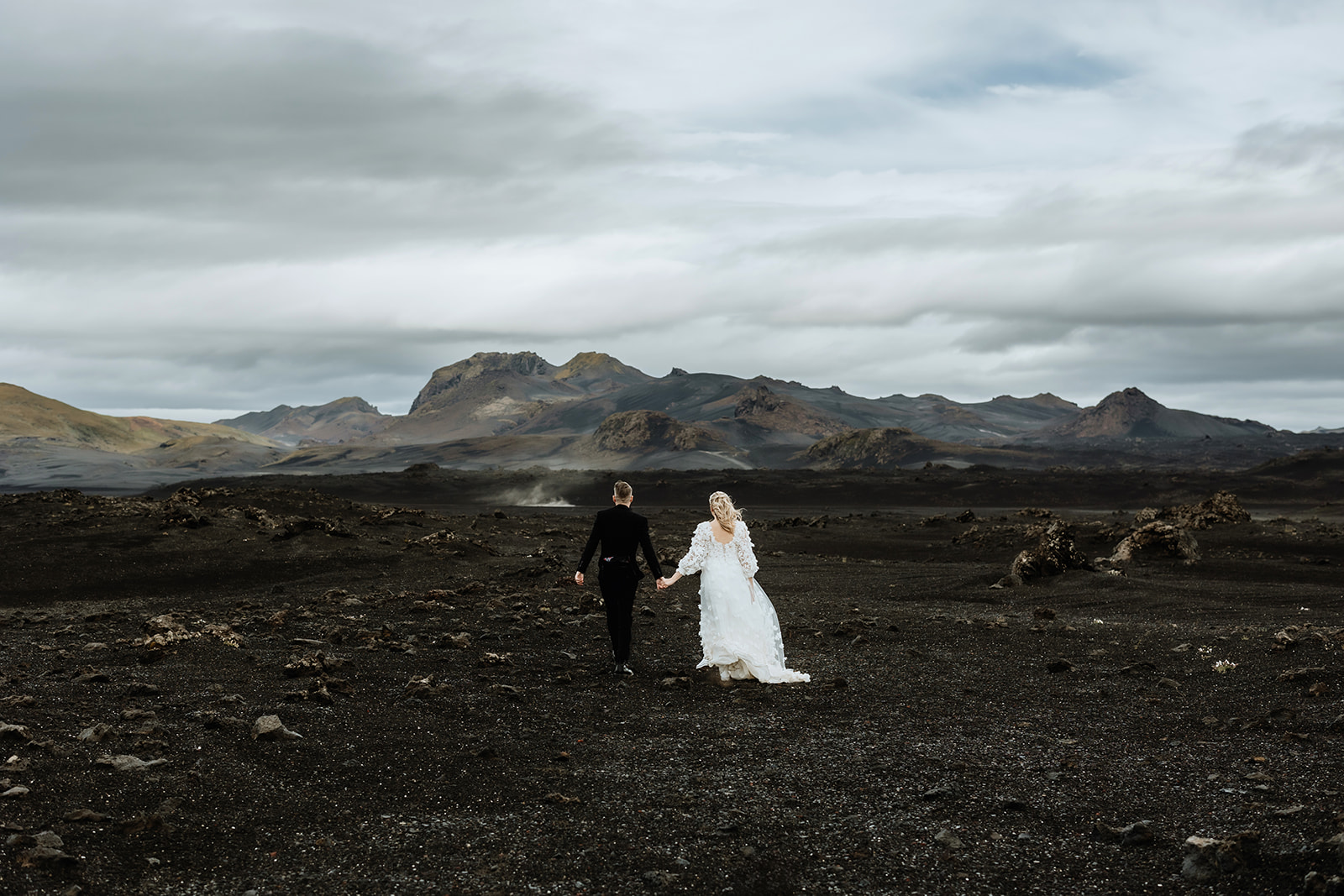 Newlywed couple holding hands and walking through the Icelandic highlands, with rolling hills and a serene landscape.