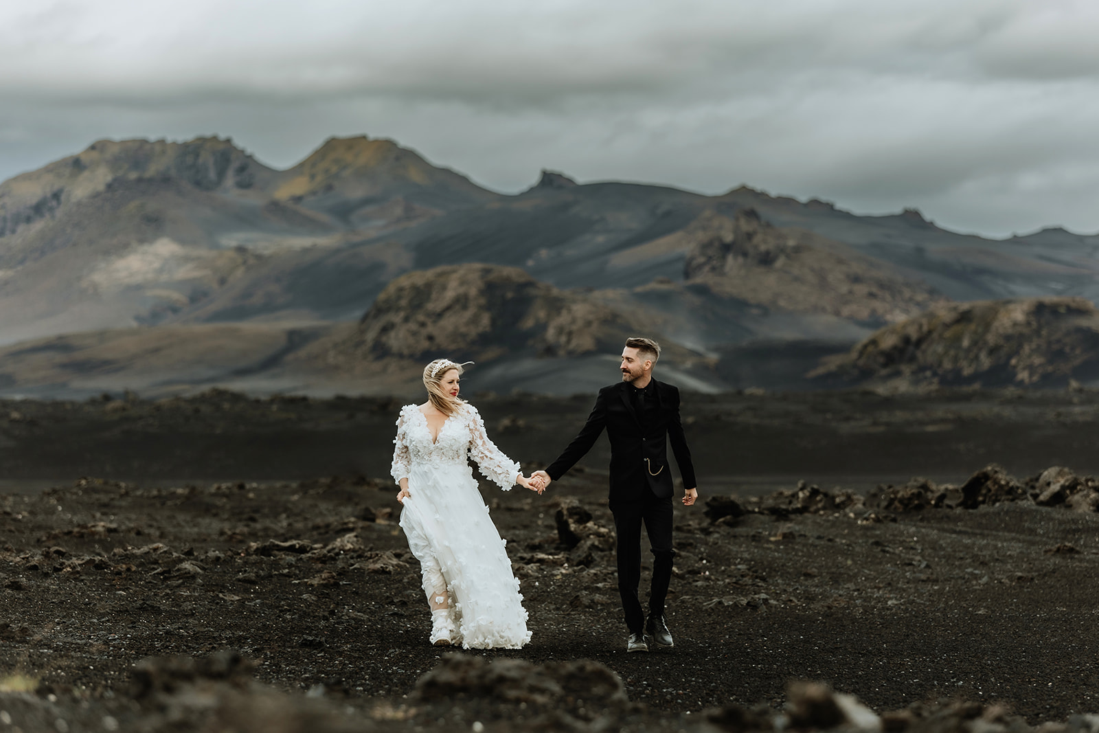 Newlywed couple holding hands and walking through the Icelandic highlands, with rolling hills and a serene landscape. 