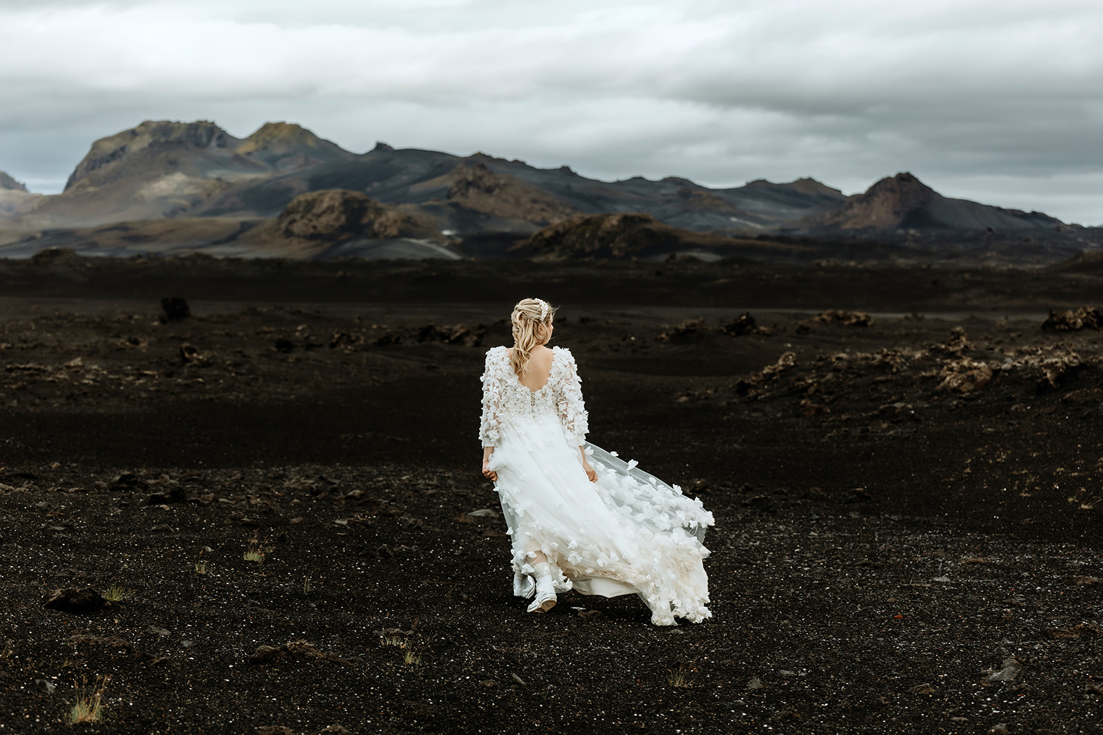 Bride in elegant wedding dress walking through dramatic Icelandic highlands, with rugged terrain and moody skies
