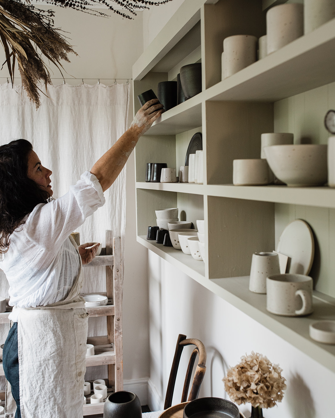 Woman reaches for ceramic jug on shelf of pottery items.