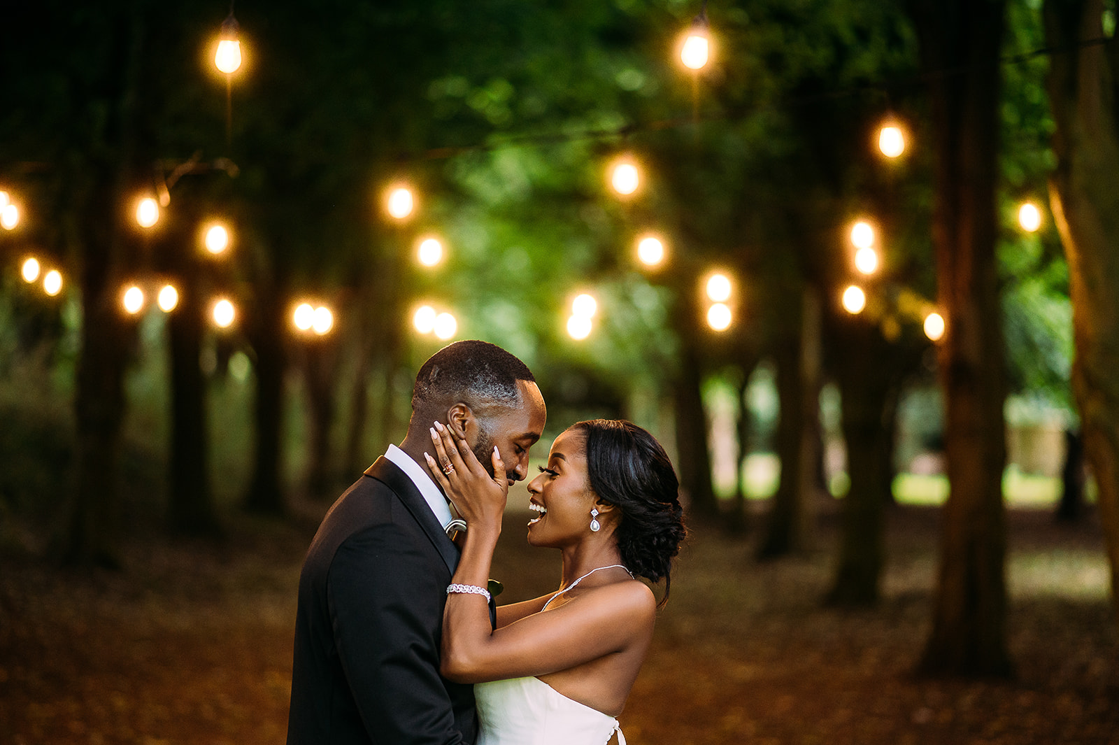 Intimate couple portrait under festoon lights on a tree lined path.