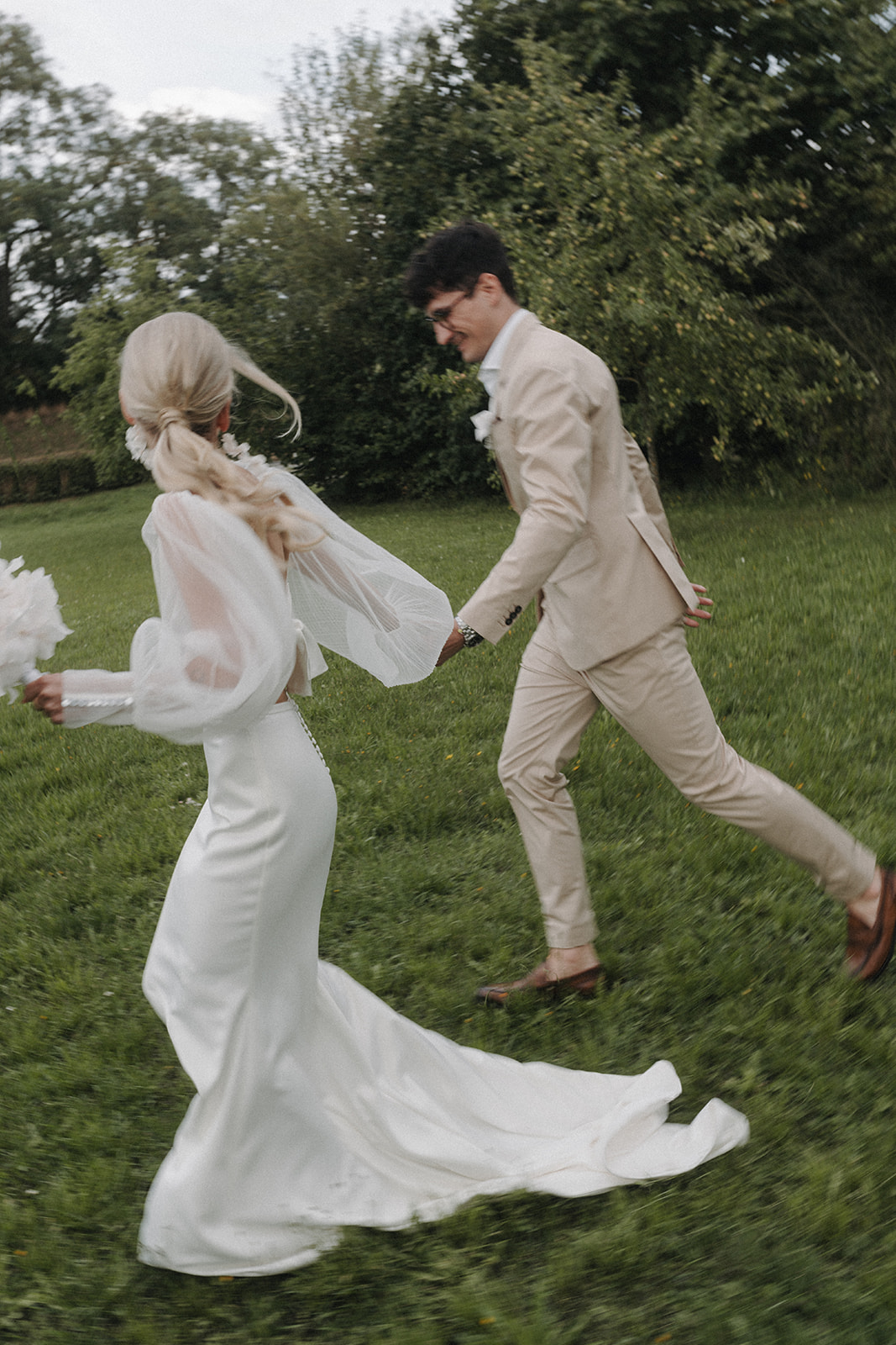 Braut und Bräutigam laufen über grüne Wiese und halten dabei ihre Hände. Sie trägt ein modern chic Kleid. 