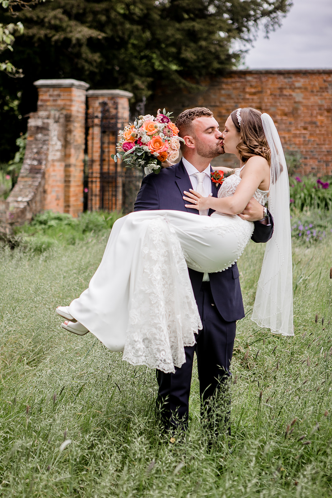 groom carrying his bride through the long grass at Blake Hall