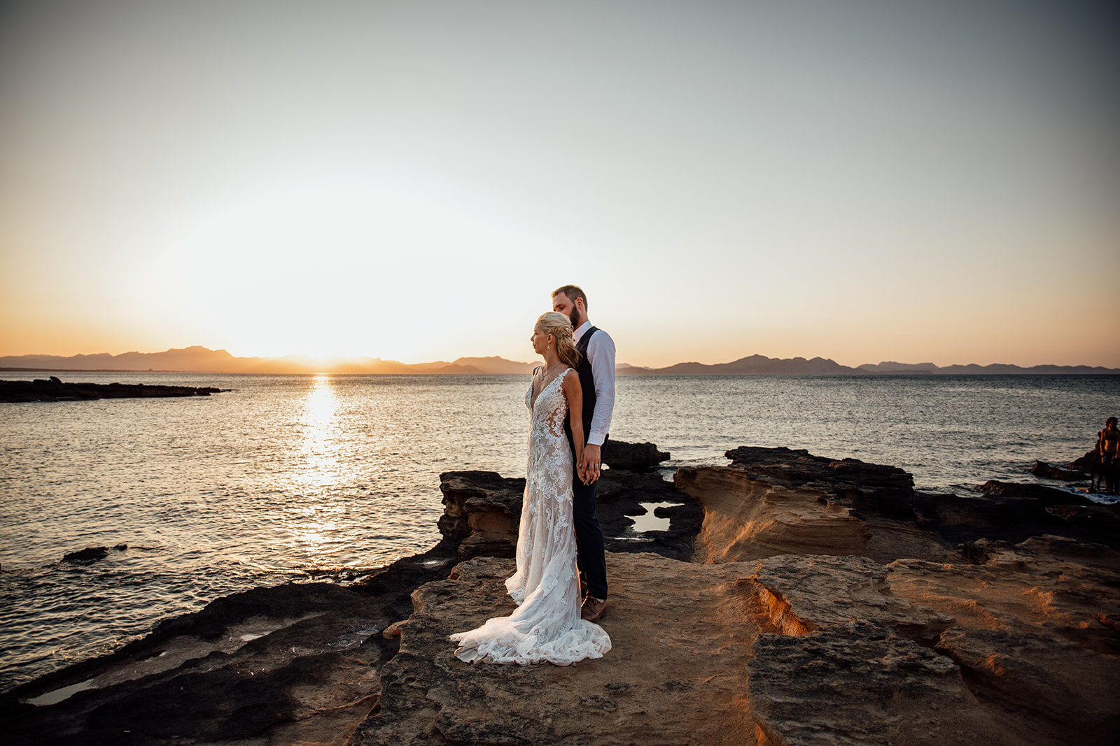 After Wedding Shooting Mallorca - Paar steht vorne am Meer, links geht die Sonne unter 