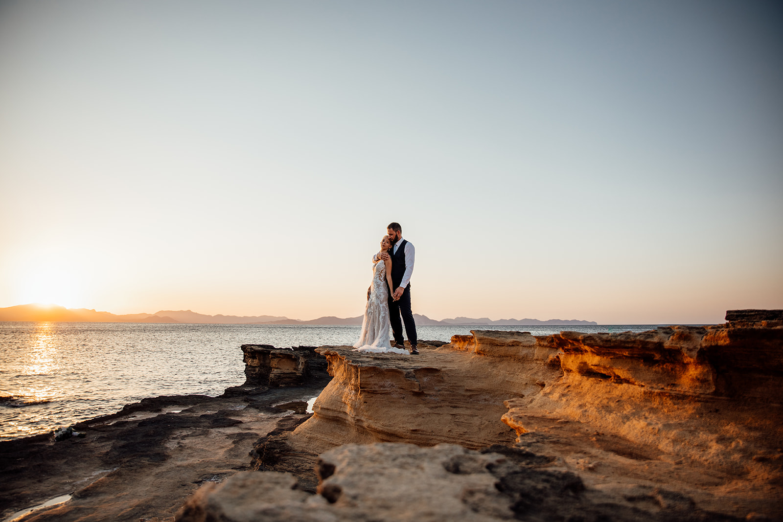 After Wedding Shooting Mallorca - Paar steht vorne am Meer, links geht die Sonne unter 