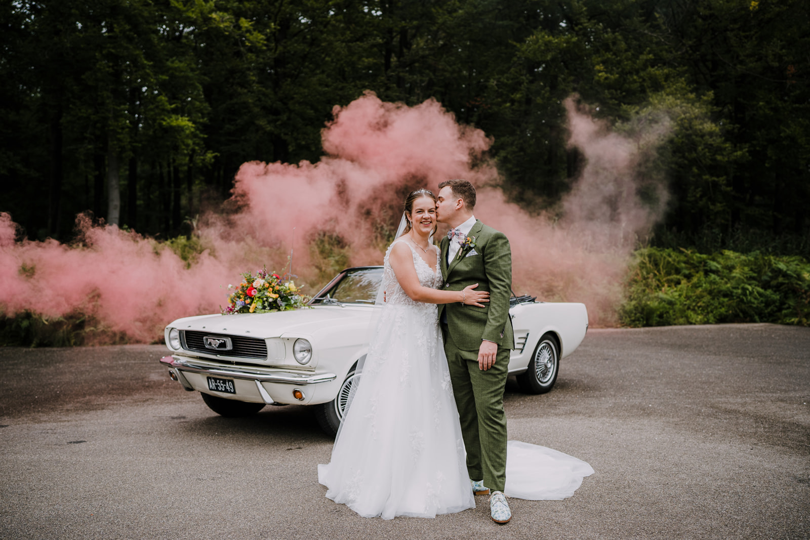 Bruidspaar poseert met Ford Mustang op Landgoed Staverden met sfeervolle roze rook flare op de achtergrond in het bos