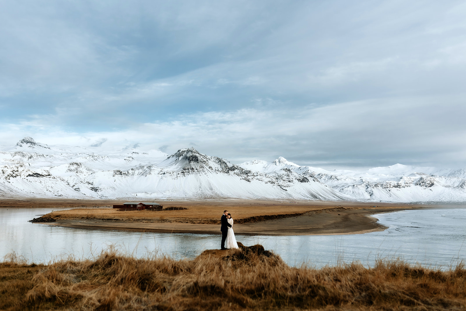 Newlywed couple is standing in Iceland surrounded by snowy mountains and breathtaking winter landscape with ocean.