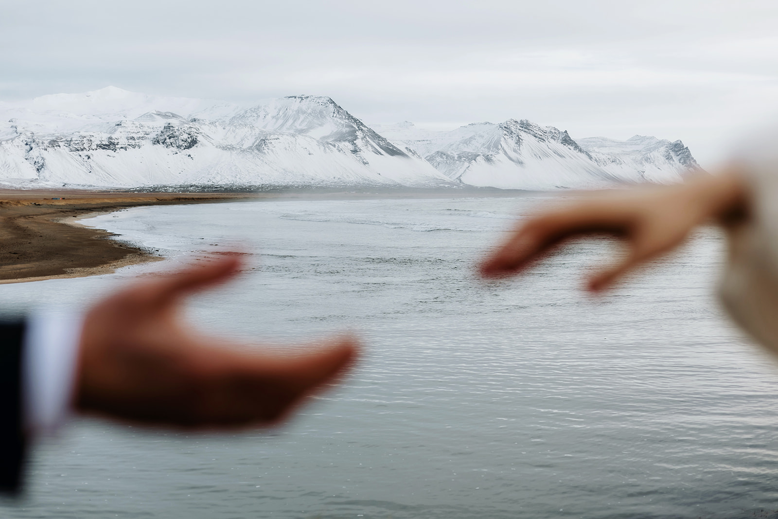 married couple hands are reaching towards each other, snowy mountains are in the back. Iceland winter elopement.