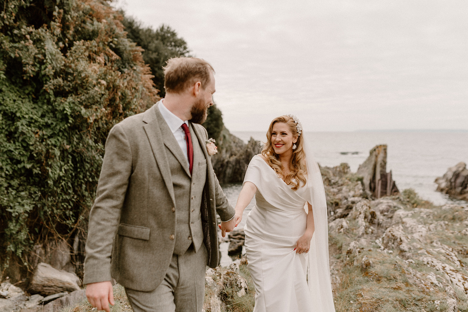 Bride and groom walk hand in hand on rocks overlooking sea at Polhawn Fort