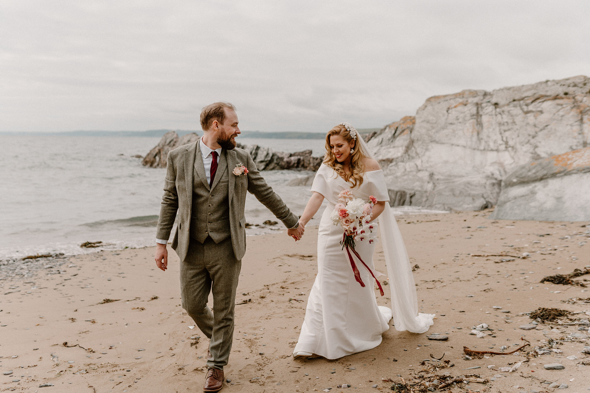 Bride and Groom walk hand in hand on sandy beach.